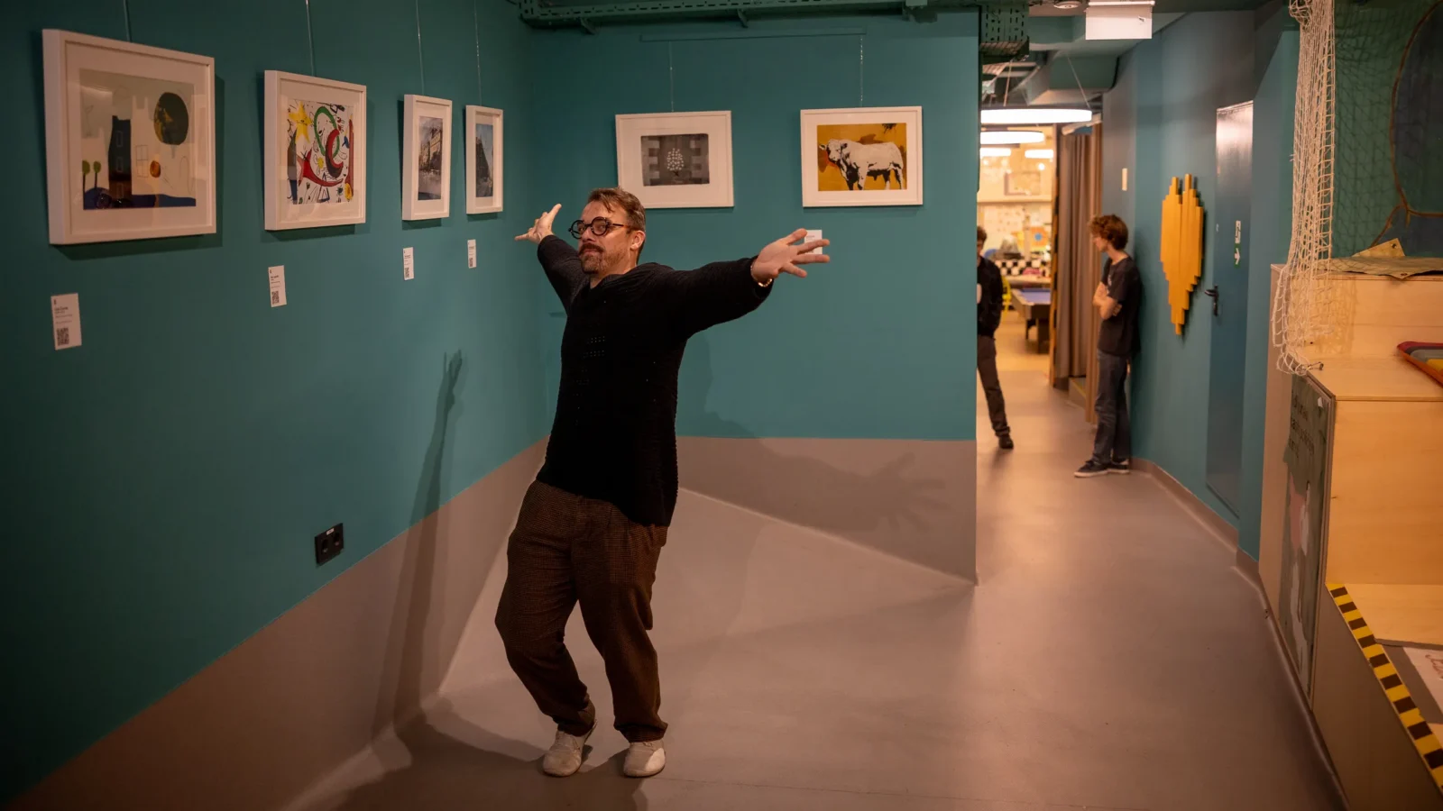 A person extending both arms in a gallery with turquoise walls and framed works.